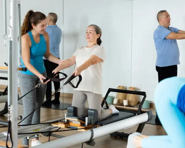 Frauen und Männer trainieren im **Reha-Sport** auf Pilatesgeräten. Eine Trainerin hilft einer älteren Frau, während ein Mann mit einem Ball übt.