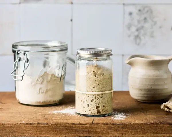 Zwei Gläser mit Sauerteig Weizen, eines mit aktivem, blasigem Sauerteig, das andere mit trockenem Ansatz. Daneben ein kleiner Krug.
