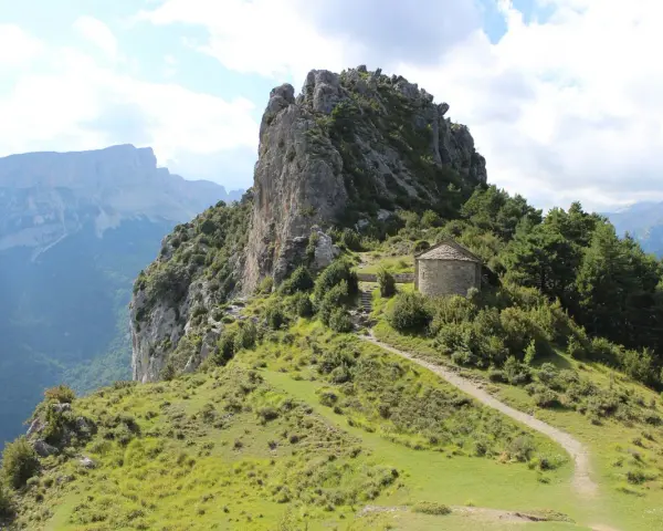 Pequeña ermita de piedra en la ruta de las ermitas, rodeada de vegetación y con imponentes montañas al fondo.