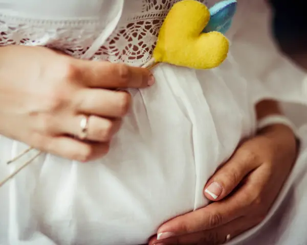 Brzuszek w białej sukience, ozdobiony serduszkiem. Ręce delikatnie go obejmują. To piękny obrazek z pierwszej wizyty u położnej w ciąży.