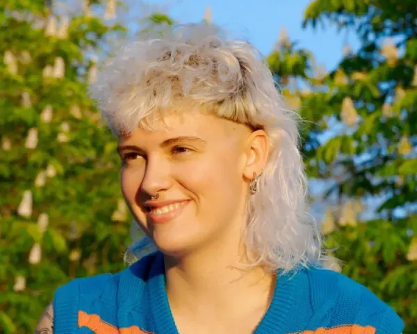 Persona sonriente con un moderno mullet rubio, aretes y un suéter azul con rayas naranjas, frente a un árbol en flor.