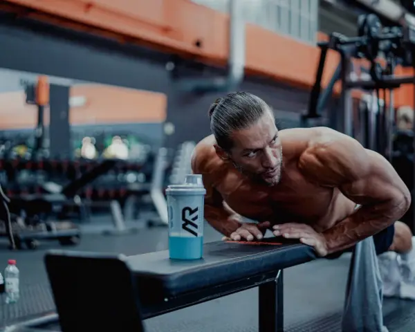 Man gör armhävningar på en bänk i gymmet, en shaker med blå dryck står bredvid. Detta är ett exempel på hur byggs muskler.