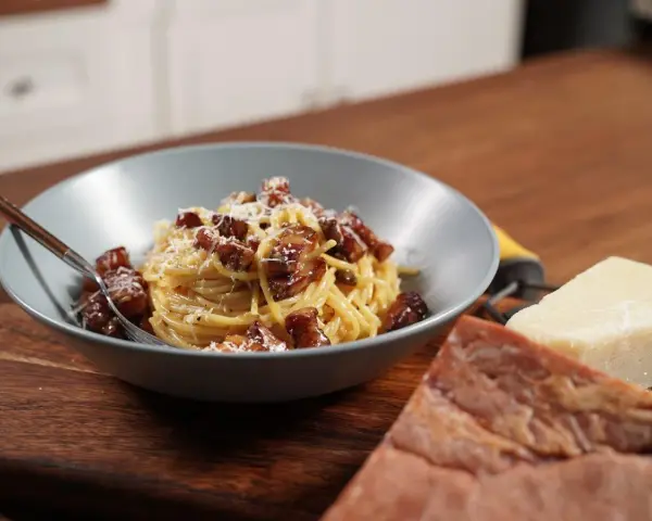 Miseczka spaghetti carbonara z boczkiem i parmezanem, obok kawałki sera i boczku.