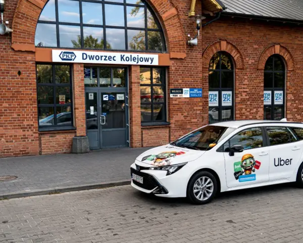 Biały samochód Ubera zaparkowany przed dworcem kolejowym. Kierowca z własnym samochodem czeka na pasażerów.