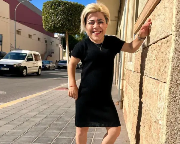 Mujer sonriente con vestido negro, apoyada en pared de piedra. Sus medidas cuerpo mujer son únicas y hermosas.