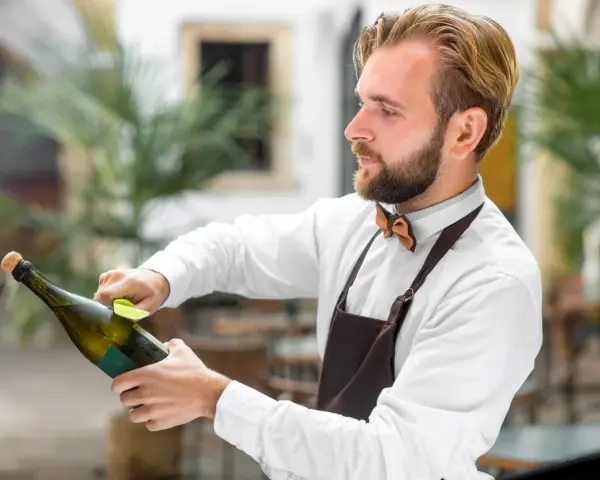 Barman w białej koszuli i brązowym fartuchu pokazuje, jak otwierać szampana, trzymając butelkę i wyciągając korek.