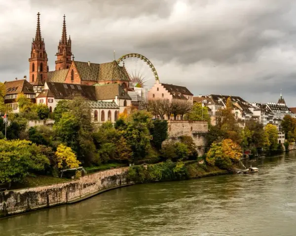 A scenic view of Basel's historic Minster and riverside buildings, perfect for a Basel itinerary. A Ferris wheel peeks out from behind the architecture.