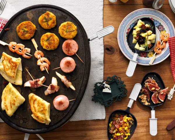 Raclette-Ideen für Kinder: Garnelen-Spieße, kleine Frikadellen, Käse-Speck-Röllchen und Crêpes auf einem Raclette-Grill.