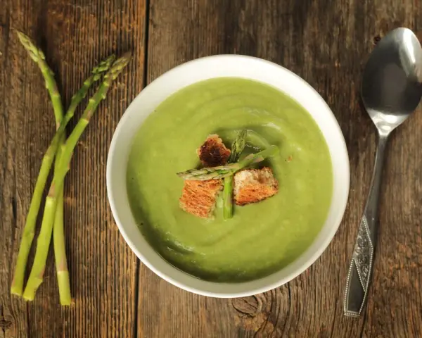 Ciotola di vellutata verde brillante, guarnita con crostini e asparagi freschi. Accanto, un mazzetto di asparagi e un cucchiaio. Un delizioso pesto di asparagi pronto da gustare.