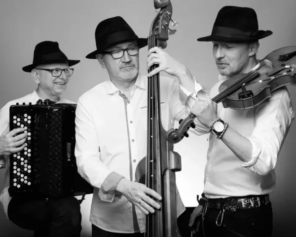 Trzech muzyków w czarnych kapeluszach grających muzykę klezmerską: akordeonista, basista i skrzypek.