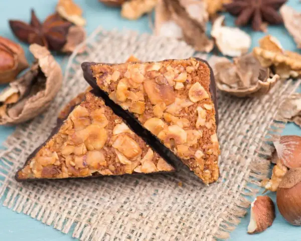 Zwei vegane Nussecken mit Schokoladenrand, bedeckt mit gehackten Nüssen, liegen auf einem Jutesack, umgeben von verschiedenen Nüssen und Sternanis.