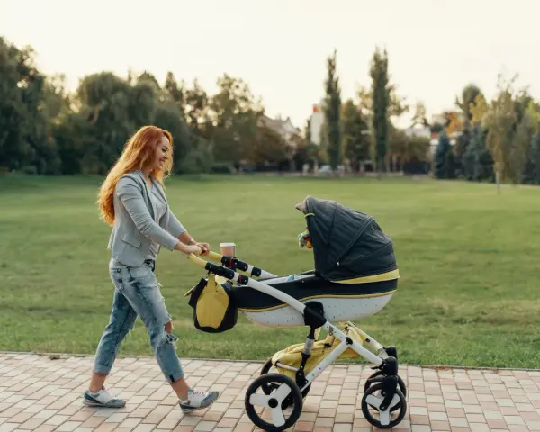 Ruda kobieta z uśmiechem pcha wózek podczas pierwszego spaceru z noworodkiem w parku.