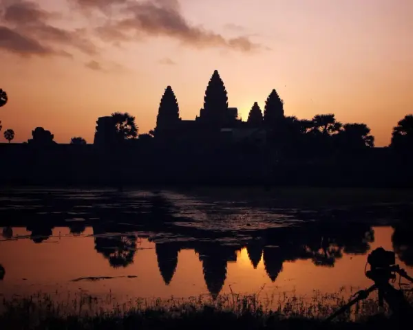 Zachód słońca nad Angkor Wat, idealne tło dla niezapomnianych kambodżańskich wakacji. Widok odbija się w wodzie, a na pierwszym planie statyw z aparatem.