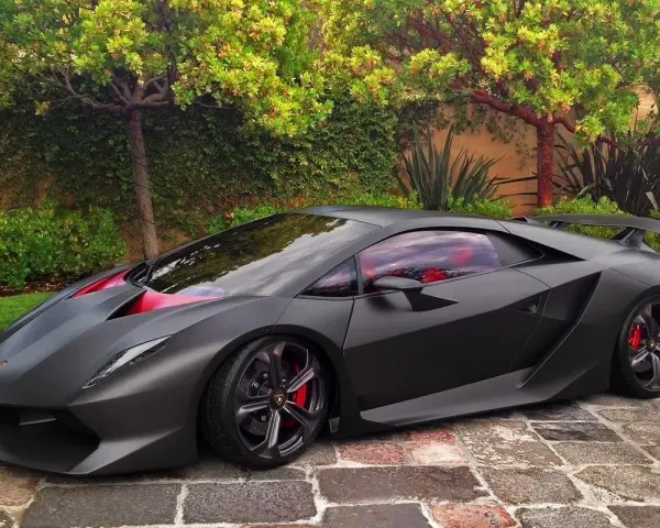 Matowy czarny Lamborghini Sesto Elemento z czerwonymi akcentami i wnętrzem, zaparkowany na brukowanej ścieżce wśród zieleni.