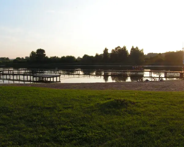 Drewniane pomosty na spokojnym jeziorze starogrodzkim, z piaszczystą plażą i zieloną trawą w tle.