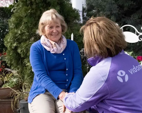Eine freundliche ältere Dame im Gespräch mit einer Pflegekraft. Deren Kleidung trägt das Logo "vidon". Ein Gutachter für den Pflegegrad könnte hier im Gespräch sein.