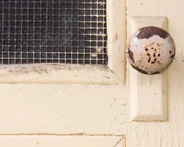 Maniglia usurata su porta interna tamburata, pronta per essere rinnovata. La rete della finestra è visibile.