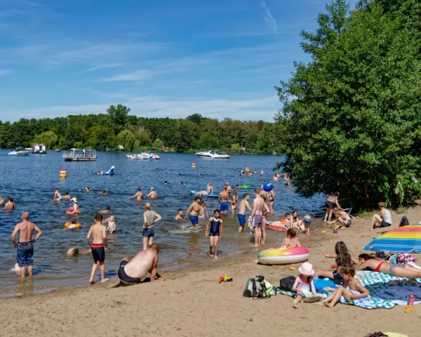 Menschen genießen das Reschensee baden an einem sonnigen Tag. Kinder planschen, Erwachsene schwimmen und entspannen am Ufer. Boote gleiten über das Wasser.