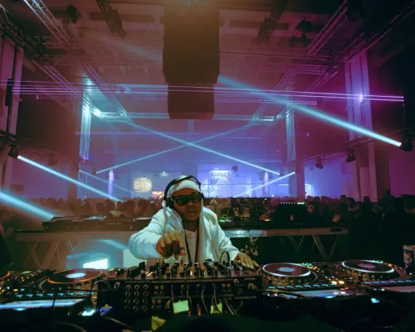 Un DJ année 2000, lunettes de soleil et bandeau, mixe sous des lasers bleus et violets devant une foule en délire.