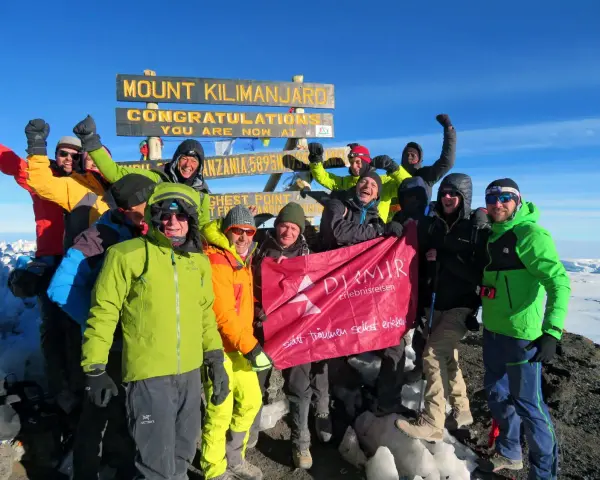 Glückliche Gruppe feiert auf dem Gipfel des Kilimandscharo. Sie haben den Gipfel erreicht und halten ein Banner hoch. Ein unvergessliches Erlebnis, den Kilimandscharo zu besteigen!