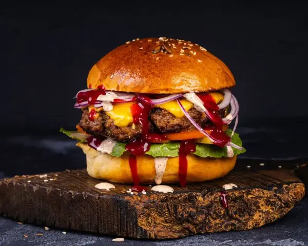 Soczyste burgery z grilla, polane sosem, z serem, cebulą i sałatą, na drewnianej desce.