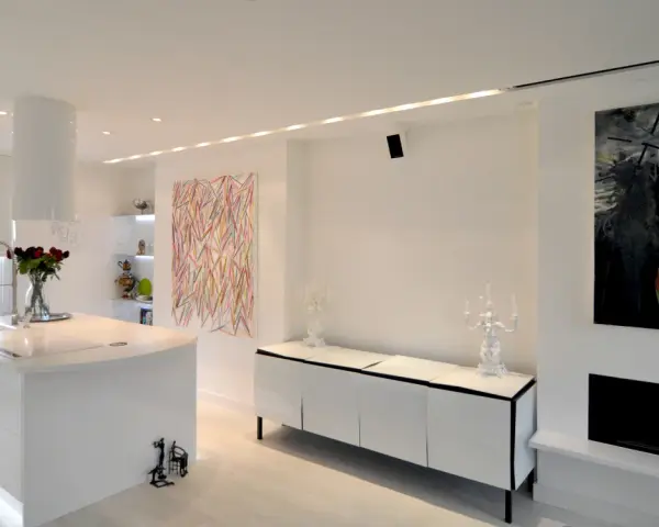 Modernt kök och vardagsrum med vita ytor, konst och spotlights som skapar en ljus och inbjudande atmosfär.