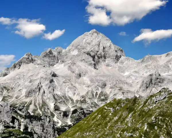 Majestätischer Triglav im Sonnenschein, bereit für die Triglav-Besteigung. Grüne Hänge und Felsen unter blauem Himmel mit Wolken.