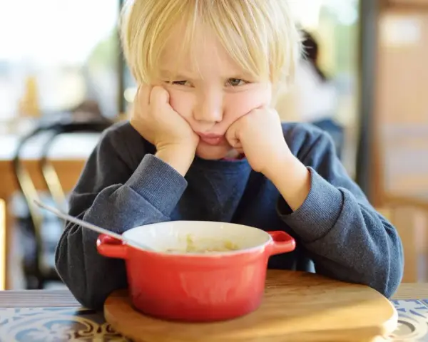 Ein Junge mit blonden Haaren stützt seinen Kopf auf die Hände und schaut lustlos auf eine Schüssel Porridge. Er isst nicht, obwohl er 10 Jahre alt ist.