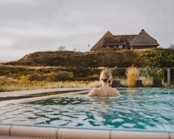 Frau genießt entspannten Wellness Tag in Schleswig-Holstein. Ein idyllischer Pool mit Blick auf Reetdachhaus und Dünenlandschaft.