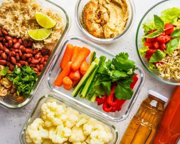 Gotowe pojemniki z jedzeniem na piknik: ryż z fasolą i pieczarkami, warzywa pokrojone w słupki, kalafior, sałatka z kiełkami i pomidorami, hummus i napoje.