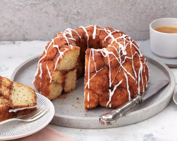 Ein köstlicher Monkey Bread mit Zimt und Zuckerguss, bereit zum Teilen. Daneben eine Tasse Kaffee und Zimtstangen.