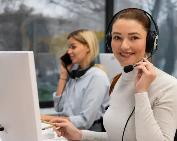 Uśmiechnięta key account manager w słuchawkach z mikrofonem pracuje przy komputerze. W tle koleżanka rozmawia przez telefon.