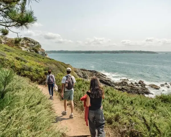 Trois randonneurs suivent un sentier côtier, profitant de la vue sur la mer. Ils avancent sur le chemin, comme dans les gr 34 étapes.