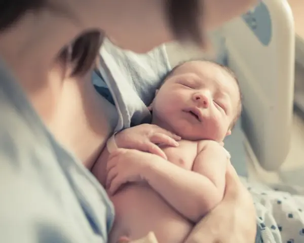 Noworodek śpi w ramionach matki, widoczna jej szyja i fragment ramienia.