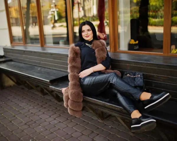 Elegancka kobieta w futrzanej kamizelce i skórzanych spodniach, zastanawia się, jakie buty do skorzanych spodni będą najlepsze. Wybrała czarne, platformowe mokasyny.