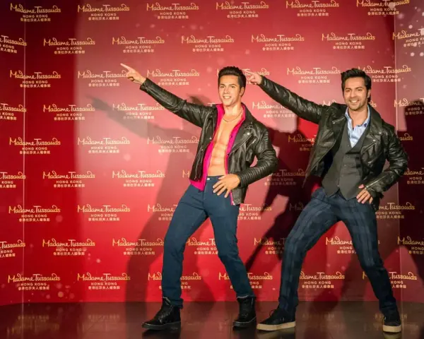 Dwie woskowe figury, jedna z odkrytym torsem, stoją na tle powtarzającego się logo Madame Tussauds w Hongkongu.