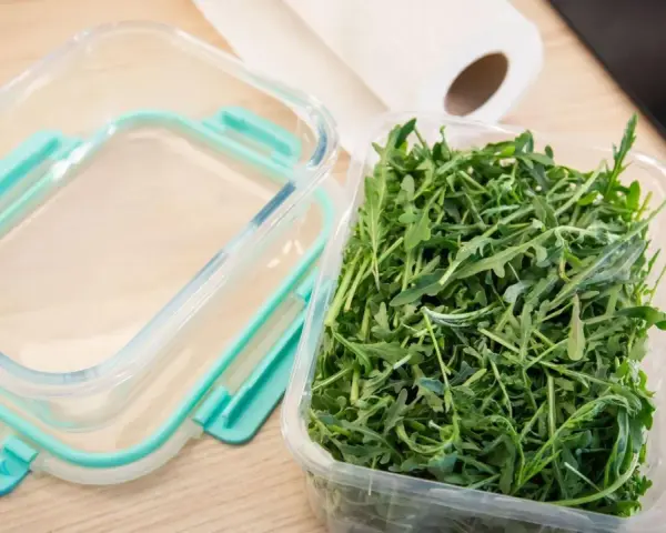 Frischer Rucola in einer Plastikschale neben einem leeren Glasbehälter. So gelingt das Rucola aufbewahren am besten.