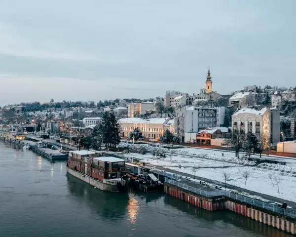 Widok na zimowe miasto nad rzeką z łodziami i ośnieżonymi budynkami.