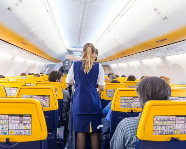 Wnętrze samolotu Ryanair, stewardessa obsługuje pasażerów. Żółte zagłówki siedzeń i pomarańczowe pasy na suficie tworzą słoneczną atmosferę.