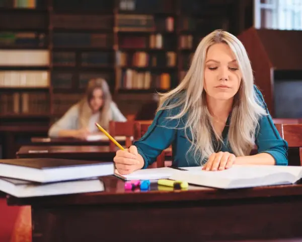 Kobieta z długimi blond włosami pisze w zeszycie, siedząc przy biurku w bibliotece.
