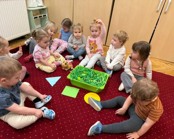 Dzieci siedzą na dywanie wokół zielonego pojemnika z zieloną trawą i jajkami.