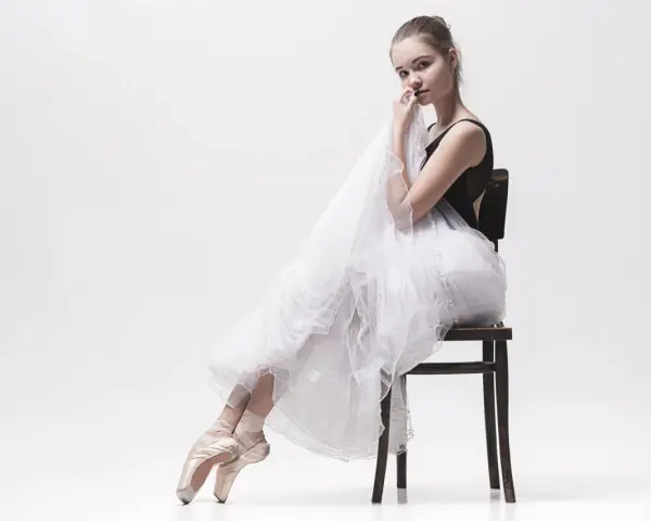 Młoda tancerka w baletkach i tiulowej spódnicy, uosobienie stylu balletcore, siedzi na krześle, trzymając w dłoniach delikatną tkaninę.