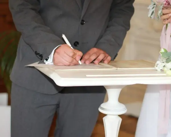 Pan młody podpisuje dokumenty, co się mówi na ślubie cywilnym? W tle panna młoda z bukietem.