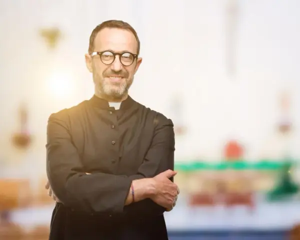 Ein lächelnder evangelischer Pfarrer mit Brille und überkreuzten Armen steht vor einem unscharfen Kircheninterieur.
