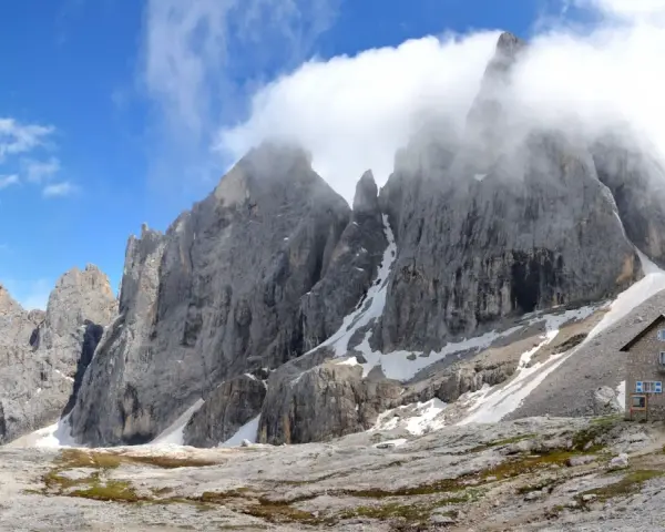 Rifugio in pietra ai piedi di imponenti vette rocciose, con neve residua e cielo azzurro. Un luogo incantevole al passo Valles.