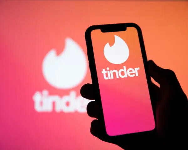 Dłoń trzyma smartfon z logo Tinder. W tle widać większe, rozmyte logo aplikacji, sugerujące nowe tinder powiadomienia.