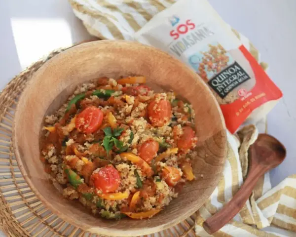 Un tazón de madera lleno de quinoa con verduras coloridas: tomates cherry, pimientos verdes y amarillos. Cerca, un paquete de quinoa integral y roja.