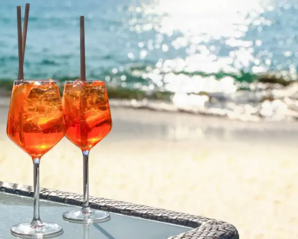 Zwei Aperol Spritz Gläser mit Eis und Orangenscheiben stehen am Strand. So trinkt man Aperol Spritz in Italien!