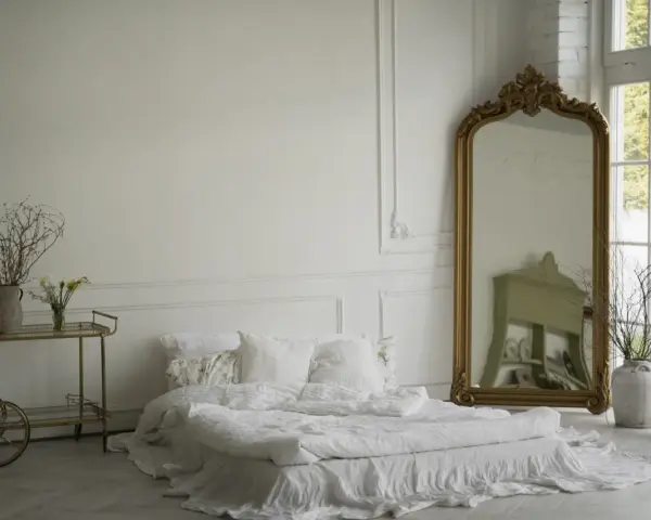 Elegancka sypialnia z łóżkiem w bieli, ozdobiona sztukaterią przypominającą lamperię angielską. Złote lustro i wózek dodają blasku.
