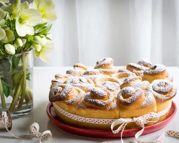 Torta di rose soffice e dorata, cosparsa di zucchero a velo, decorata con un nastro a cuoricini. Accanto, un vaso con fiori bianchi.
